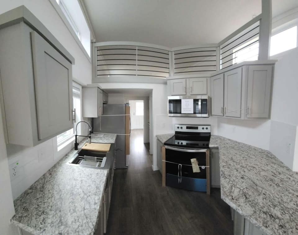 Modern kitchen with light gray cabinets, marble countertops, and stainless steel appliances. Dark wood floors and bright, upper windows enhance the space.