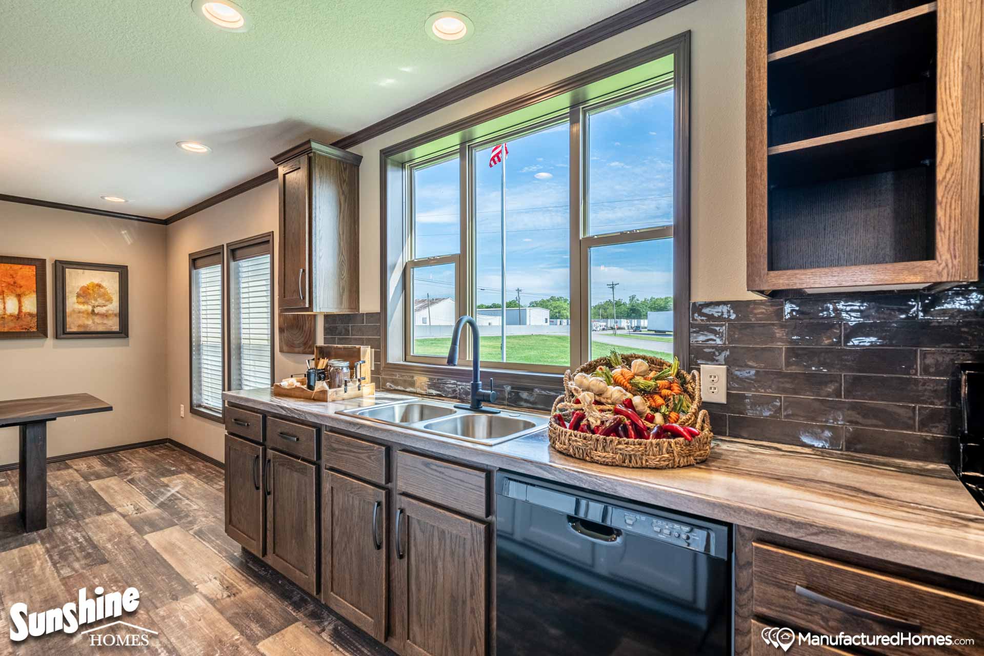 Modern kitchen with dark wood cabinets, dual sinks, and a view of a green lawn through a large window. A basket of peppers adds color to the serene space.