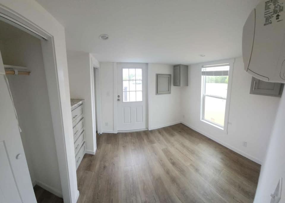 Small room with white walls and light wood floors, featuring a closed white door and a large window. There's a narrow closet with shelves on the left. The space feels bright and minimalistic.