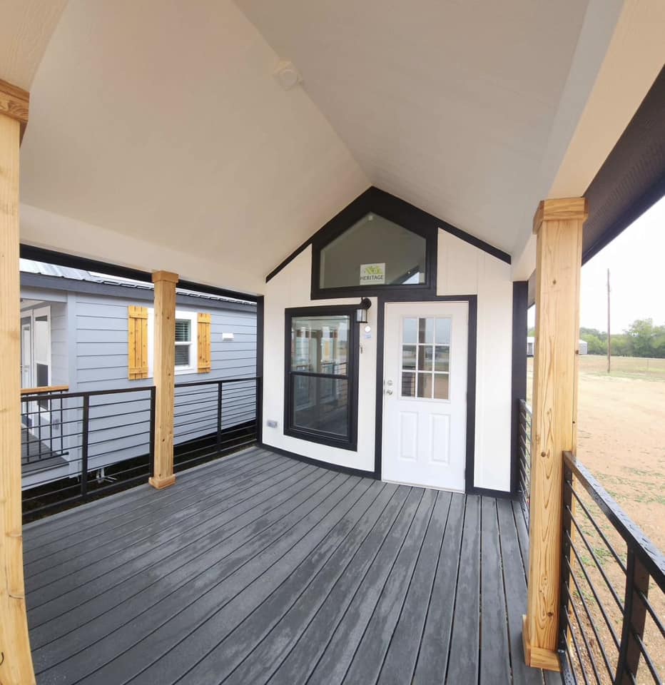 A modern porch with wooden posts and dark gray decking. It features a small white house with a gabled roof, glass door, and large window, conveying a cozy, inviting feel.