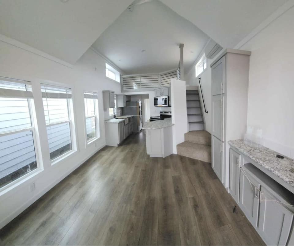 Bright open-plan kitchen with modern white and gray cabinetry, wood flooring, and large windows. Stairs lead to a loft area, creating an airy feel.