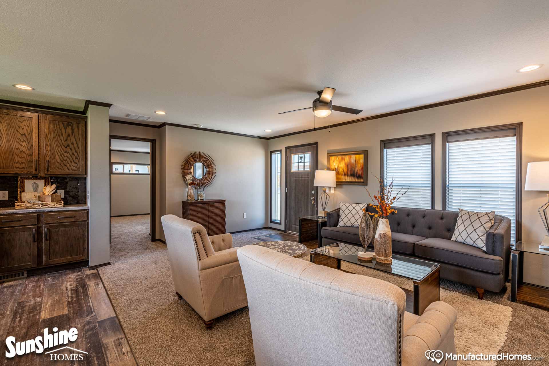 Cozy living room with beige walls and carpet, featuring a gray couch, two cream armchairs, coffee table, modern lamps, and decorative plants. Warm atmosphere.