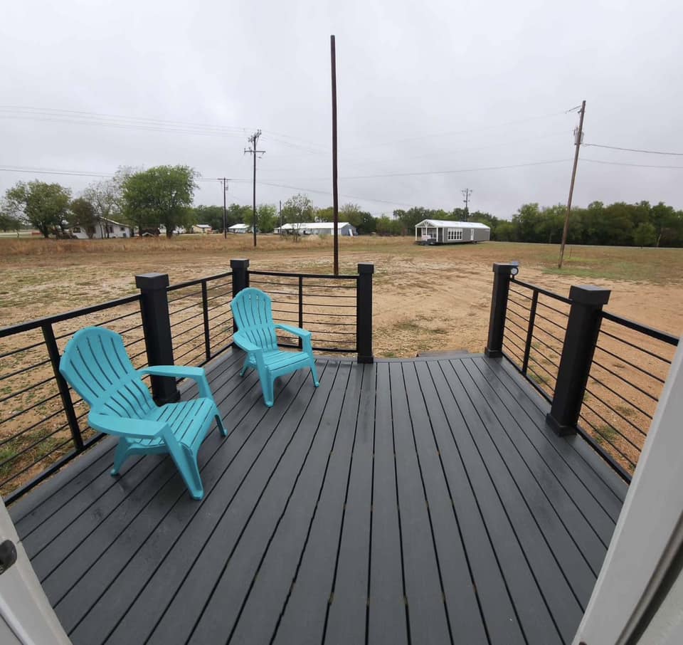 A gray wooden deck with two teal chairs overlooks a barren yard on a cloudy day. Sparse trees and a white shed are visible in the background.