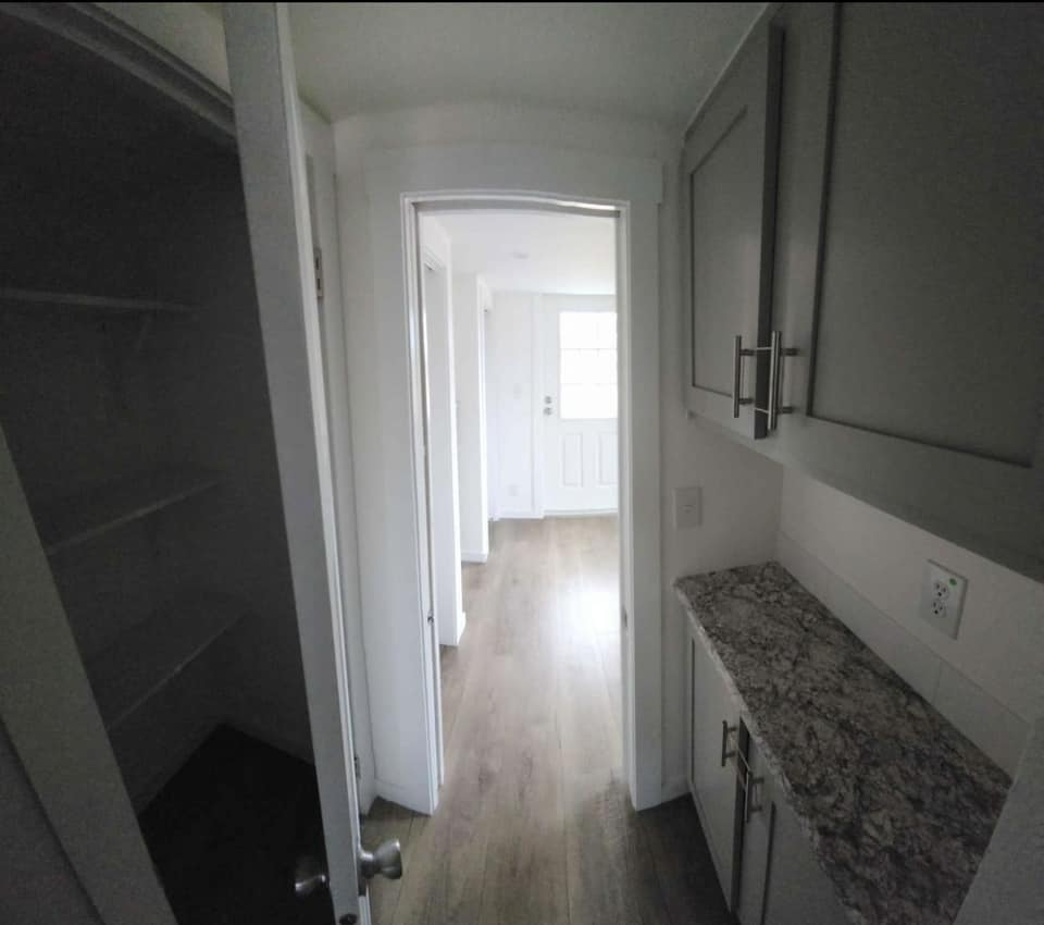 Narrow hallway with light wood flooring, white walls, and a granite counter on the right. Open closet door on the left, leading to a lit room.
