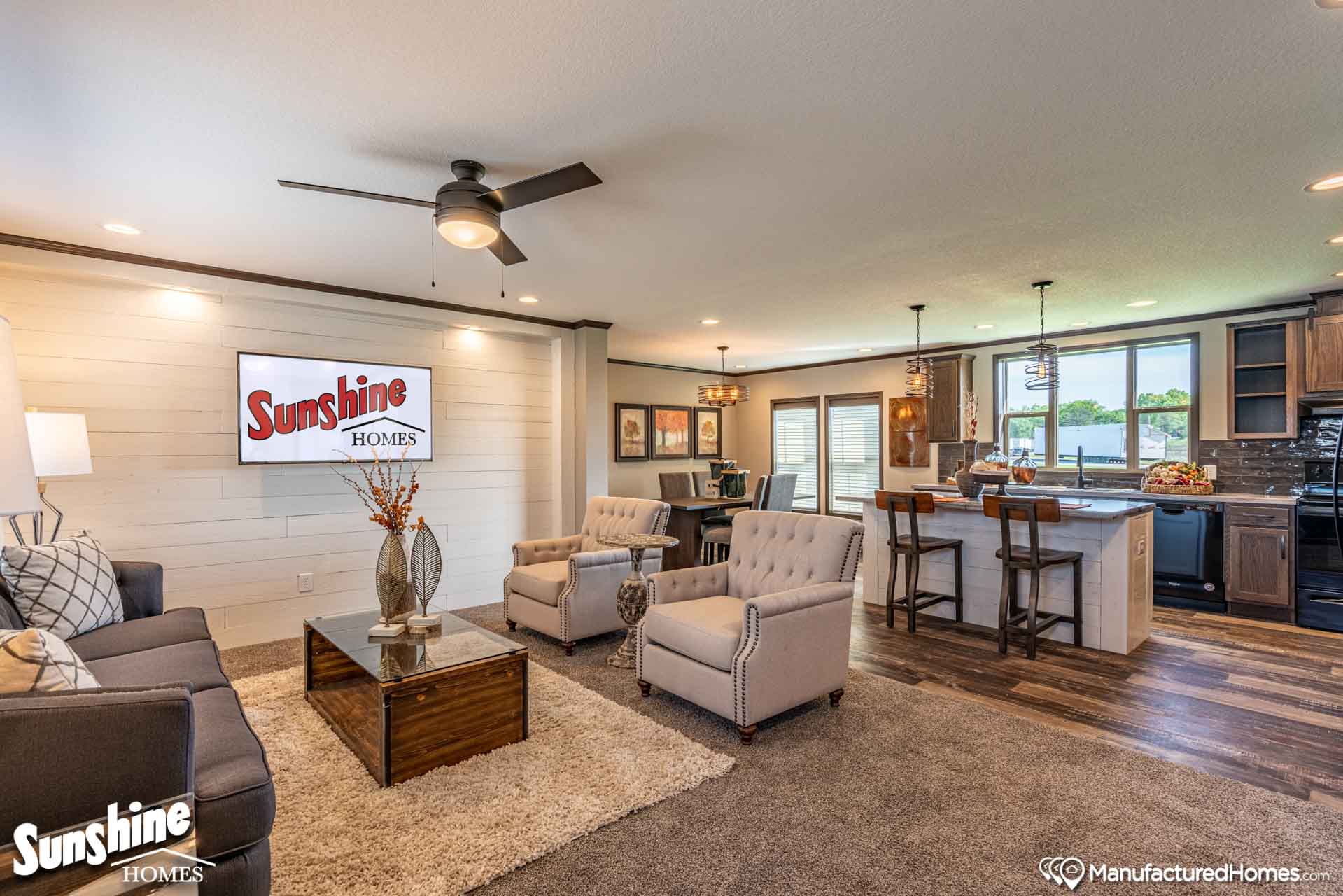 A modern living room with plush beige chairs, a dark sofa, and a stone coffee table. An open-plan kitchen with bar stools is visible. Warm lighting creates a cozy atmosphere.