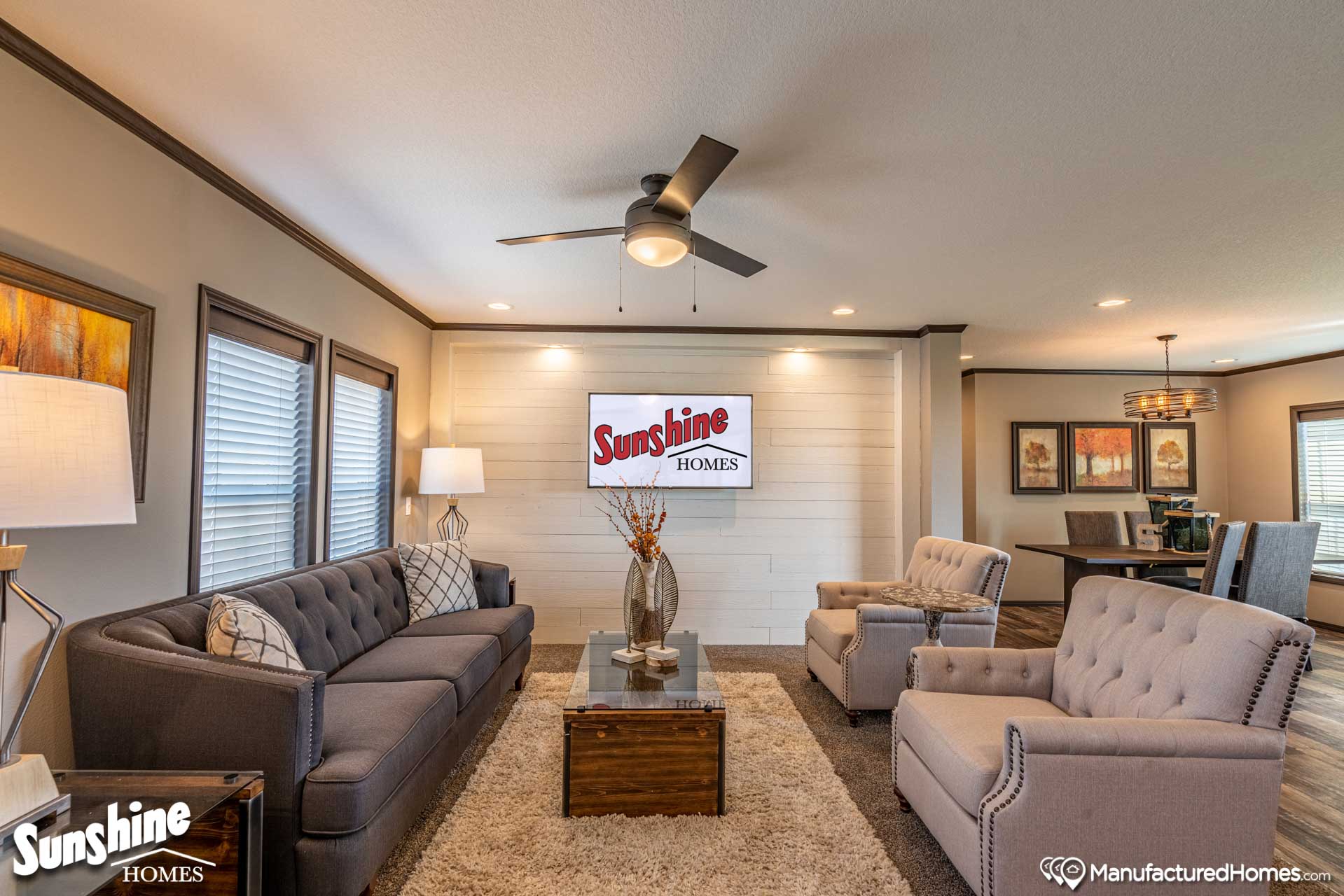 Bright, modern living room with gray sofas and armchairs, a ceiling fan, wall art, and a stylish rug. A dining area with artwork is visible in the background.