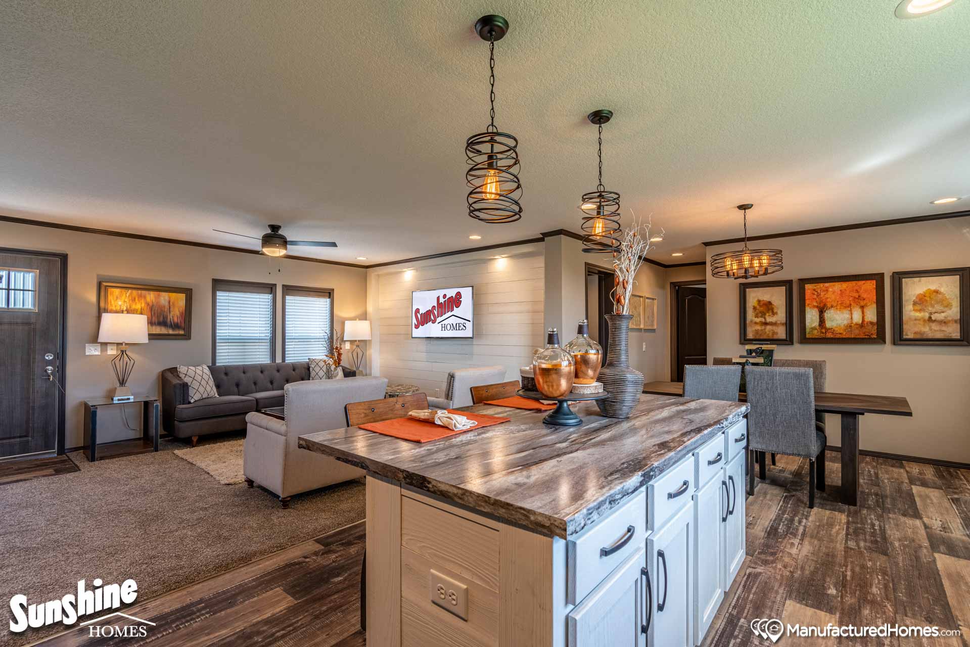 Stylish open-plan living space with a rustic kitchen island, cozy gray sofa, and warm lighting. Dining area features autumn-themed artwork, creating a welcoming atmosphere.