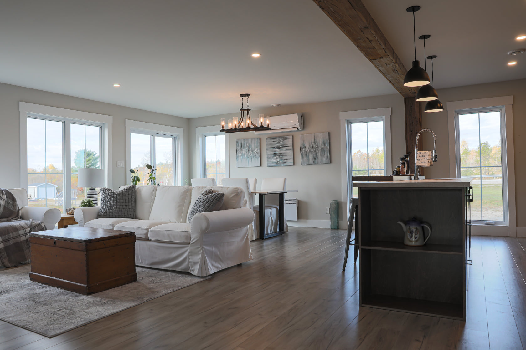 Spacious living room with modern, neutral tones. Features white sofas, a wooden coffee table, a kitchen island, large windows, and stylish lighting.