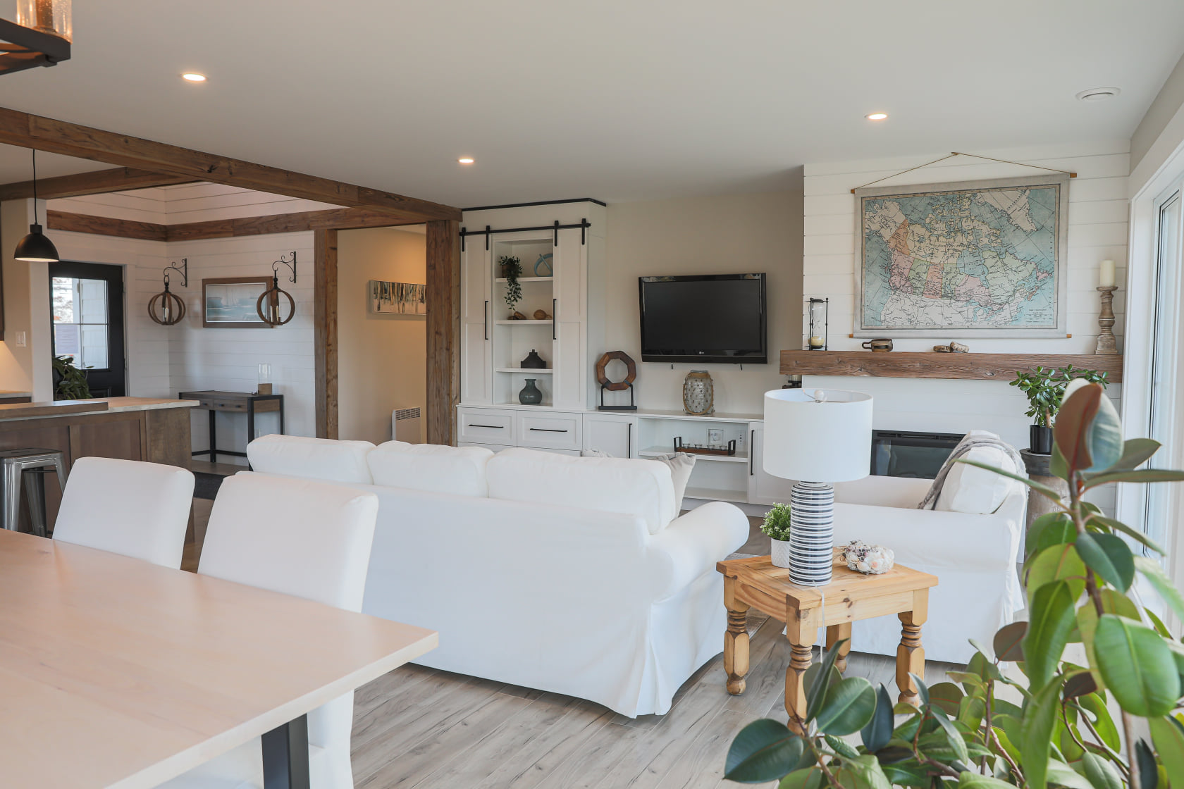Modern living room with white sofas, wooden beams, and large windows. A map hangs above a fireplace. Bright, airy, and cozy atmosphere.