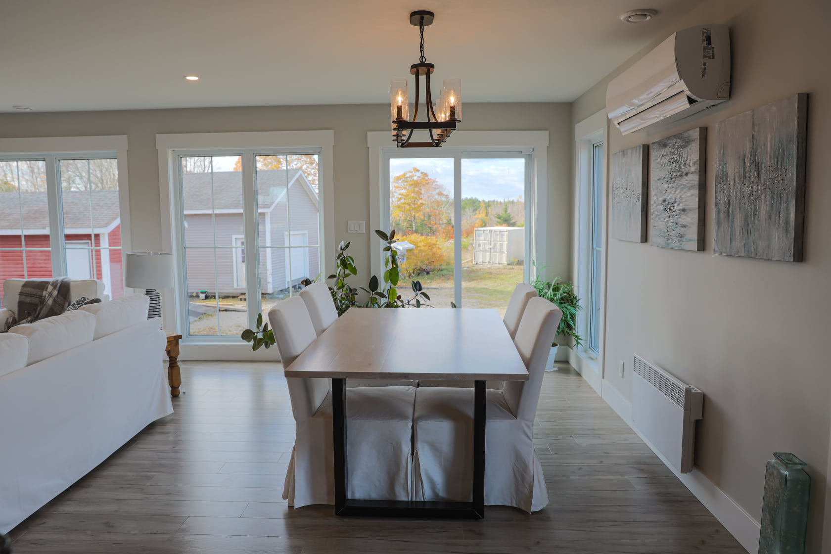 A cozy dining area with a wooden table and white chairs, under a rustic chandelier. Large windows show a fall landscape, adding warmth and light.