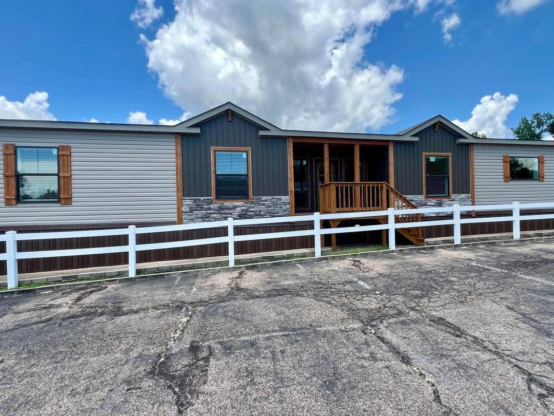 Single-story house with a dark grey facade, wooden accents, and stone detail. White fence in front and a cloudy blue sky above, creating a peaceful atmosphere.