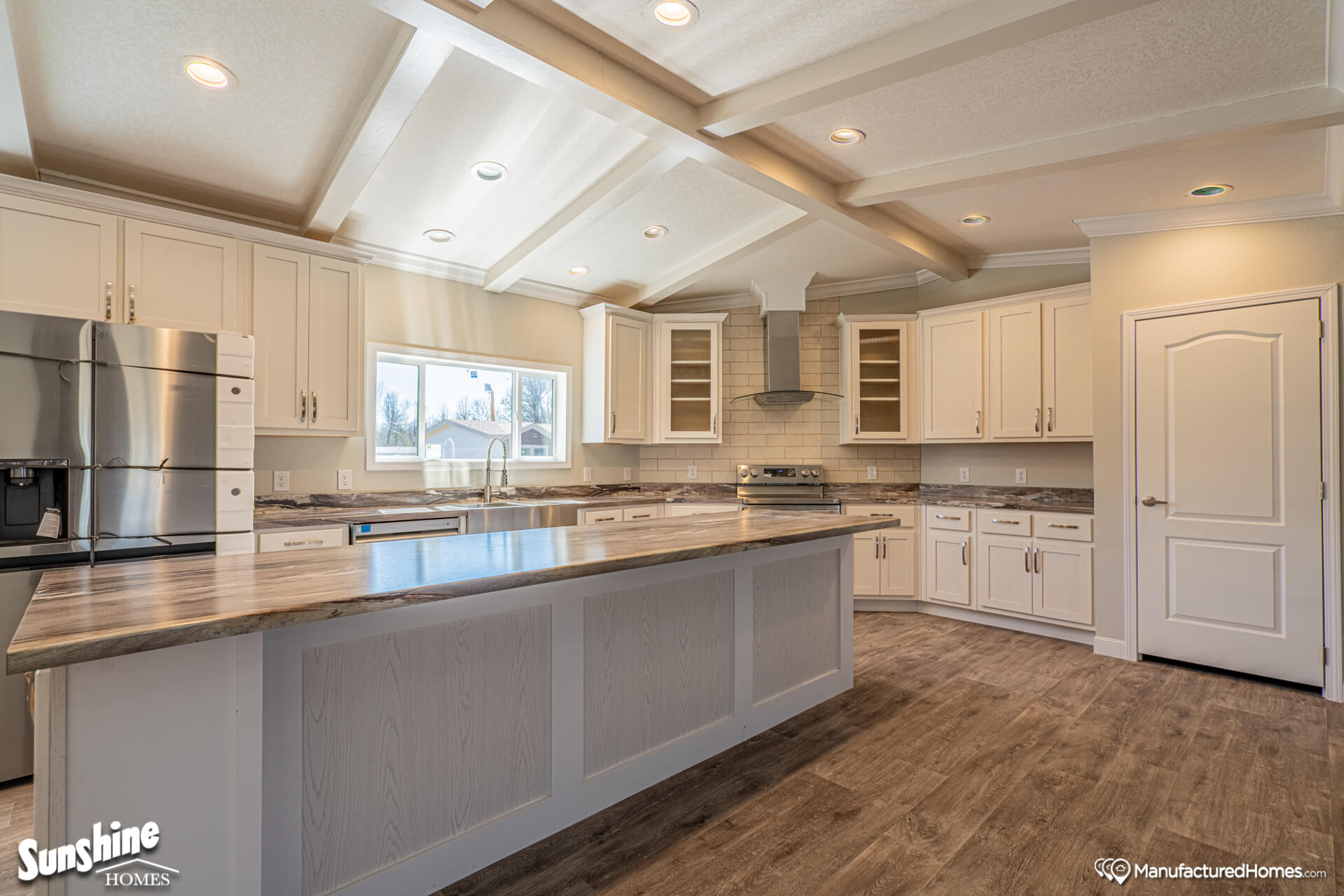 Spacious kitchen with a large island, light wood cabinets, and stainless steel appliances. Bright lighting from ceiling fixtures and a window.