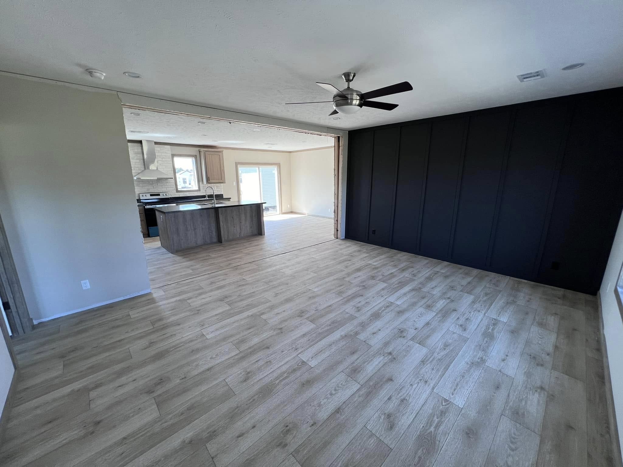 Spacious living room with light wood flooring, a ceiling fan, and a dark accent wall. Open view to a kitchen with an island and large sliding doors.