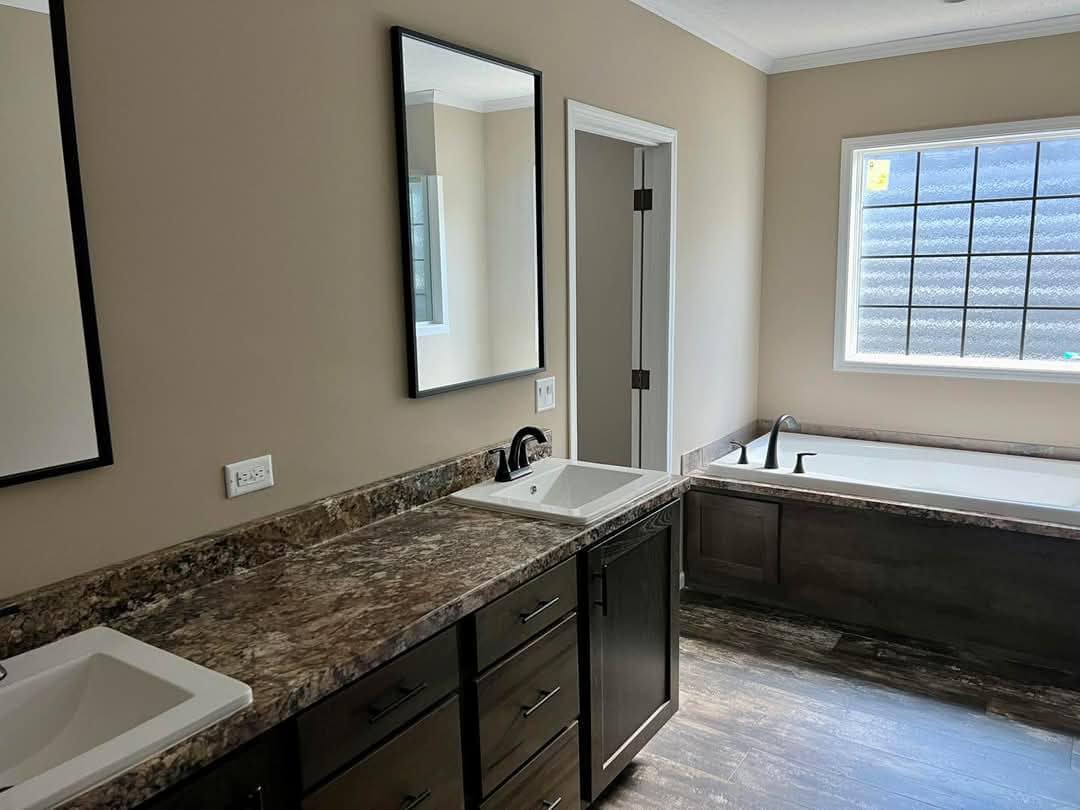 Modern bathroom with beige walls, featuring a marble double-sink vanity, framed mirrors, and a large bathtub by a window with privacy glass.