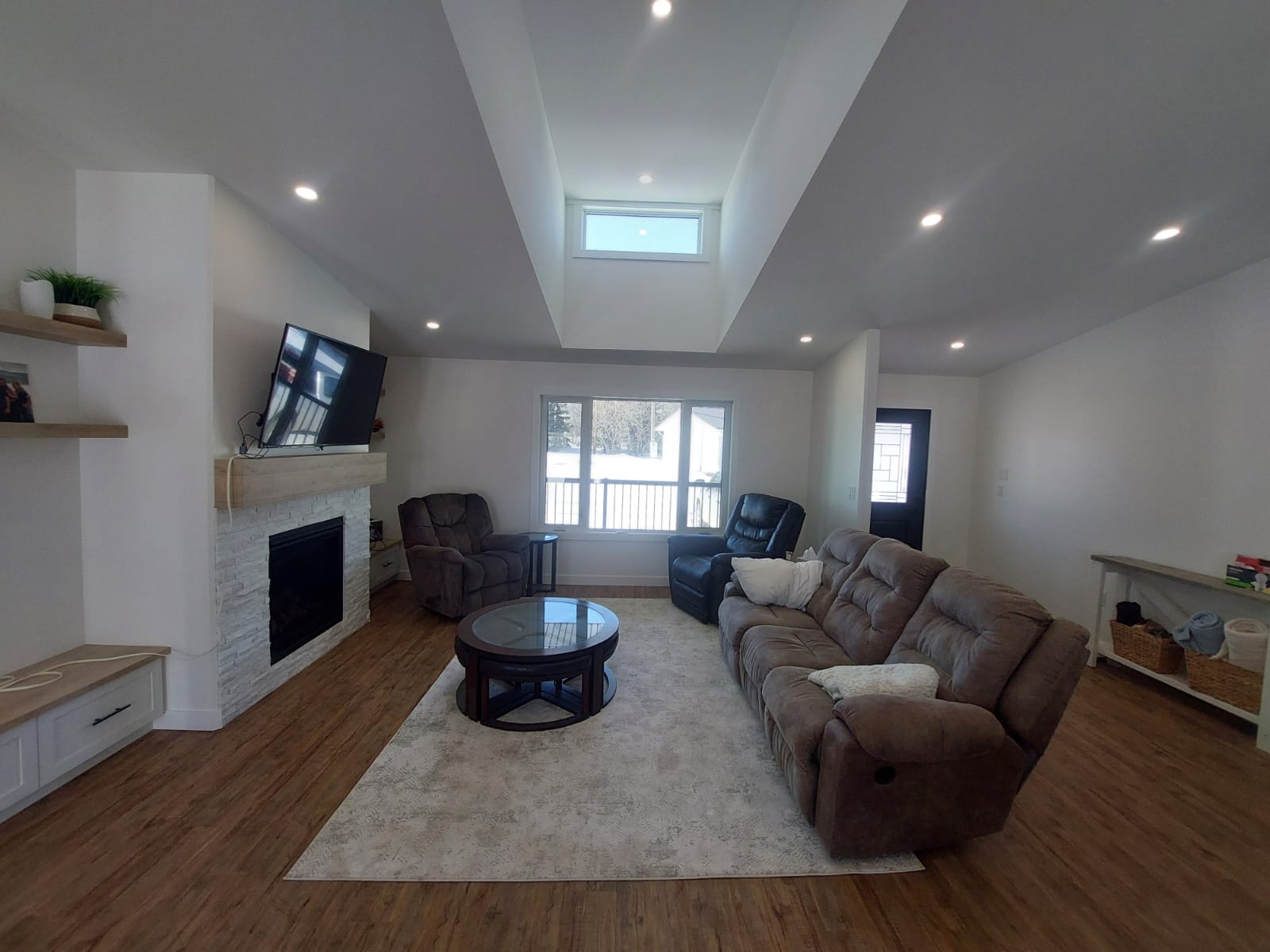 A cozy living room with brown sofas on a cream rug, a fireplace with a mounted TV, shelves with decor, and a skylight. Bright, inviting atmosphere.