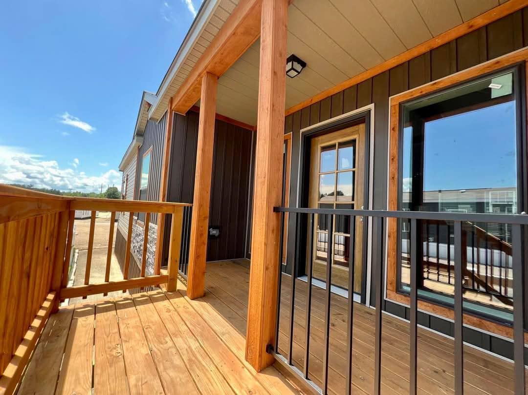 Wooden porch with railing and pillars on a sunny day, attached to a modern building with glass doors and windows, conveying a welcoming atmosphere.
