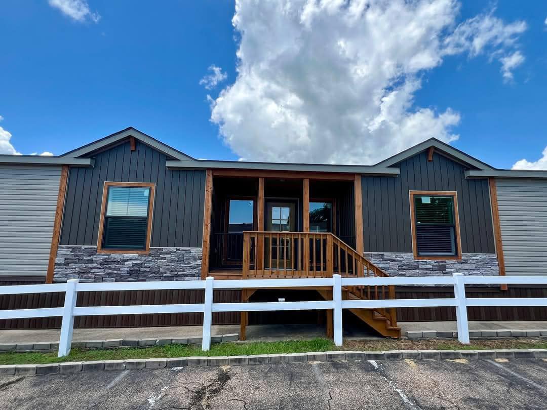 A modern modular home with dark paneling, a stone accent front, and a cozy porch is set against a bright blue sky with fluffy clouds. A white fence adds contrast.
