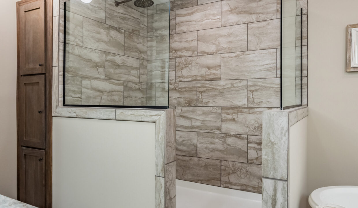 Modern bathroom shower with beige stone tile walls and glass enclosure. Adjacent is a wooden cabinet. The space feels clean and minimalist.