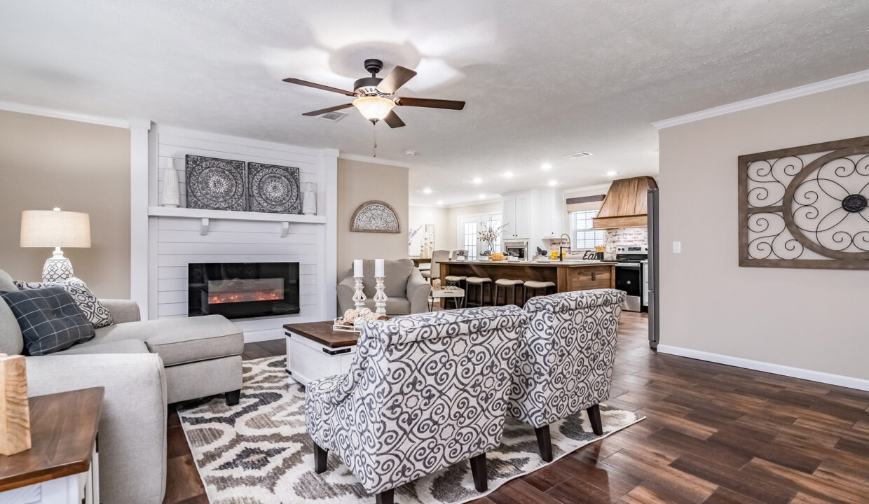 Modern, cozy living room with a fireplace, beige walls, and wood flooring. Patterned armchairs face a beige sofa. An open kitchen is visible in the background.