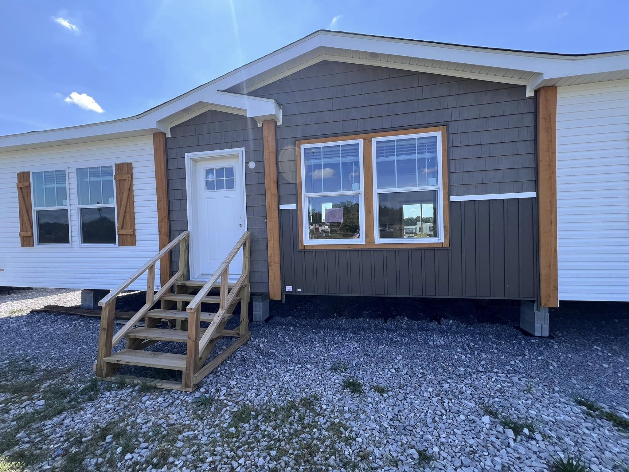 A small, single-story house with brown paneling, white siding, and wooden shutters. Wooden stairs lead to a white door. The sky is clear and sunny.