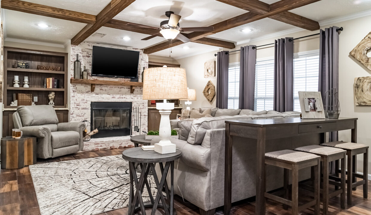 Cozy living room with a gray sofa, reclining chair, and brick fireplace under wooden beams. A large rug and table lamp add warmth and comfort.