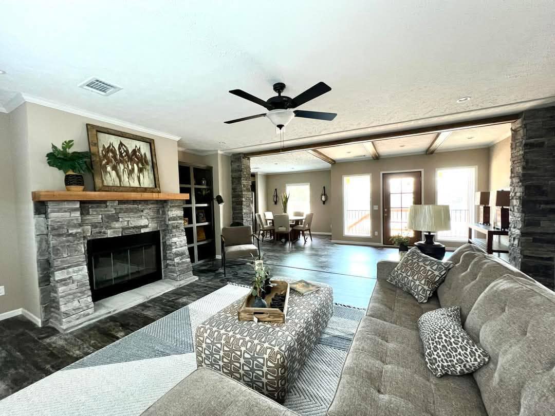 Spacious living room with a stone fireplace, gray couch, and patterned pillows. Open layout leads to a dining area with large windows and ample light.