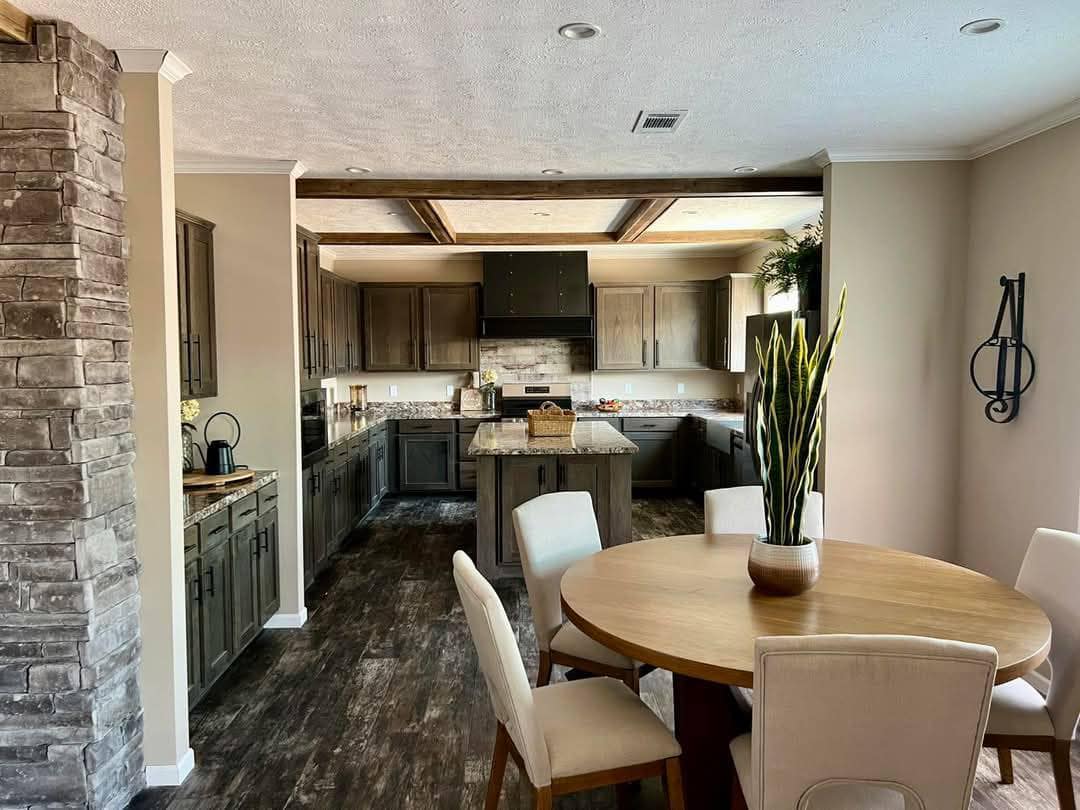 Warm and inviting kitchen and dining area with dark wood cabinets, stone accent wall, and round wooden table with white chairs. A potted plant adds a fresh touch.