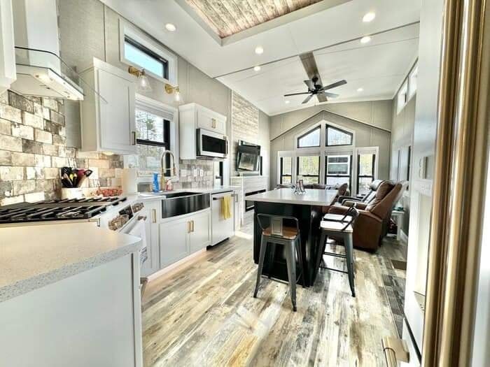 Modern kitchen and living area with white cabinets, wooden floors, and a vaulted ceiling. Features a central island, bar stools, and large windows. Cozy and bright atmosphere.