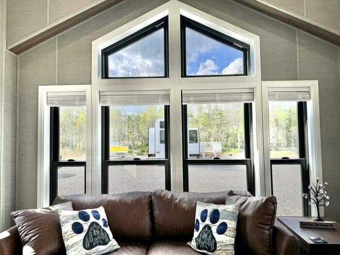 Bright living room with large geometric windows showcasing a scenic outdoor view. A cozy brown leather sofa features cushions with paw prints.