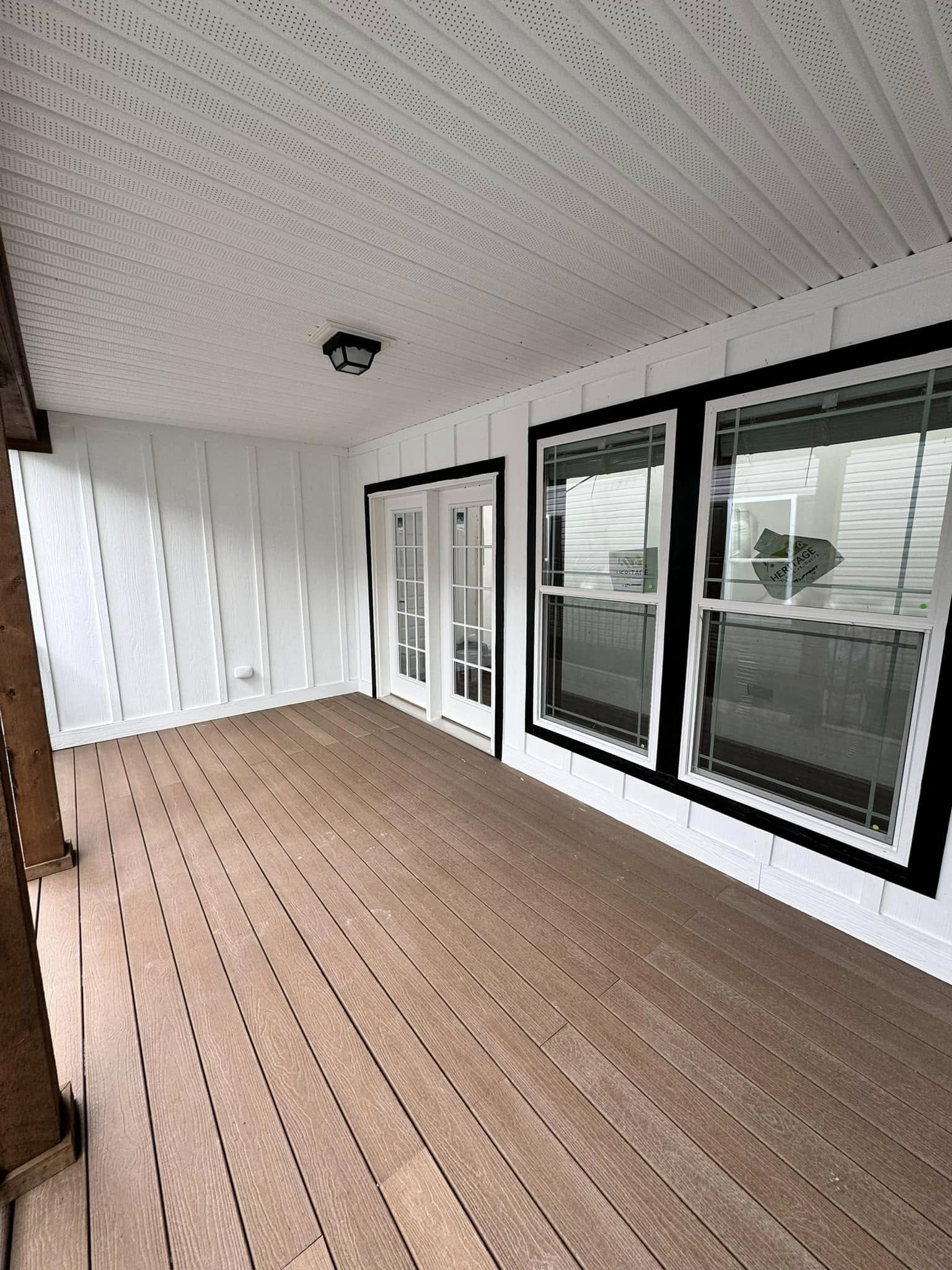 A covered patio with light brown wooden decking and white paneled walls. Glass doors and large windows framed in black create a bright and open feel.