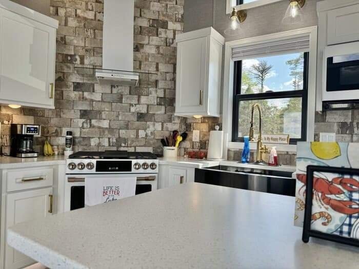 Bright, modern kitchen and living area with white cabinets, stainless steel appliances, and rustic wood floor. Cozy recliners face large, sunlit windows.