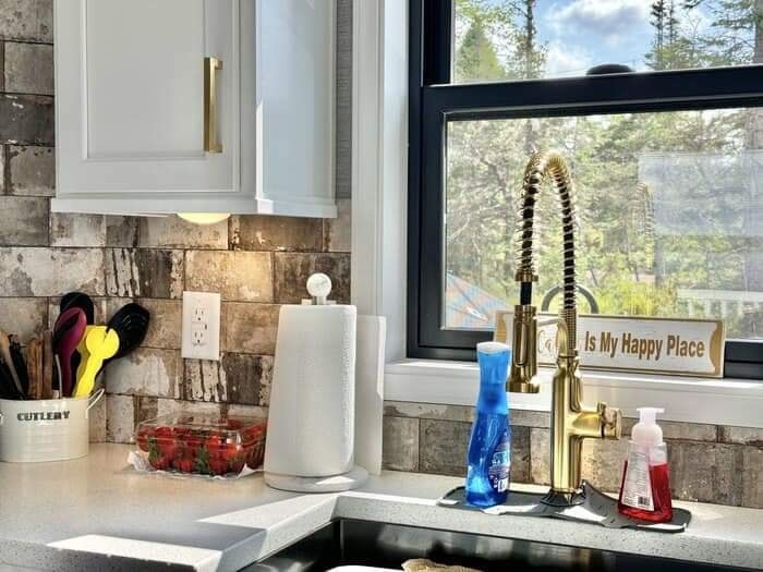 A cozy kitchen corner featuring a shiny brass faucet, soap bottles, paper towels, and strawberries. Outside the window, trees and blue sky are visible.