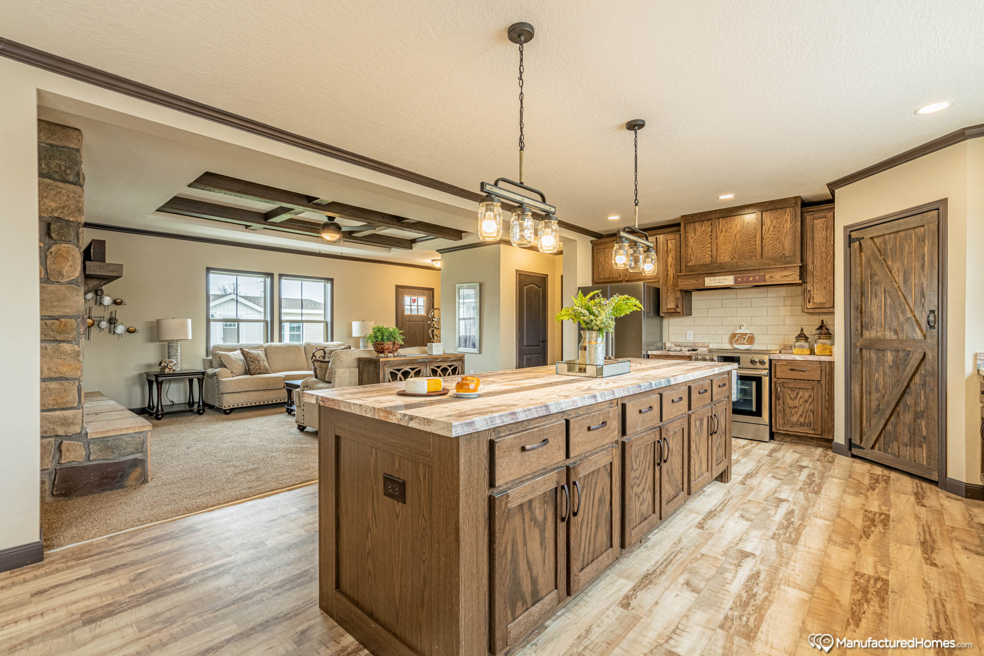 Modern kitchen with rustic wooden cabinetry, a large island, pendant lights, and stainless steel appliances. Open layout leads to a cozy living room.