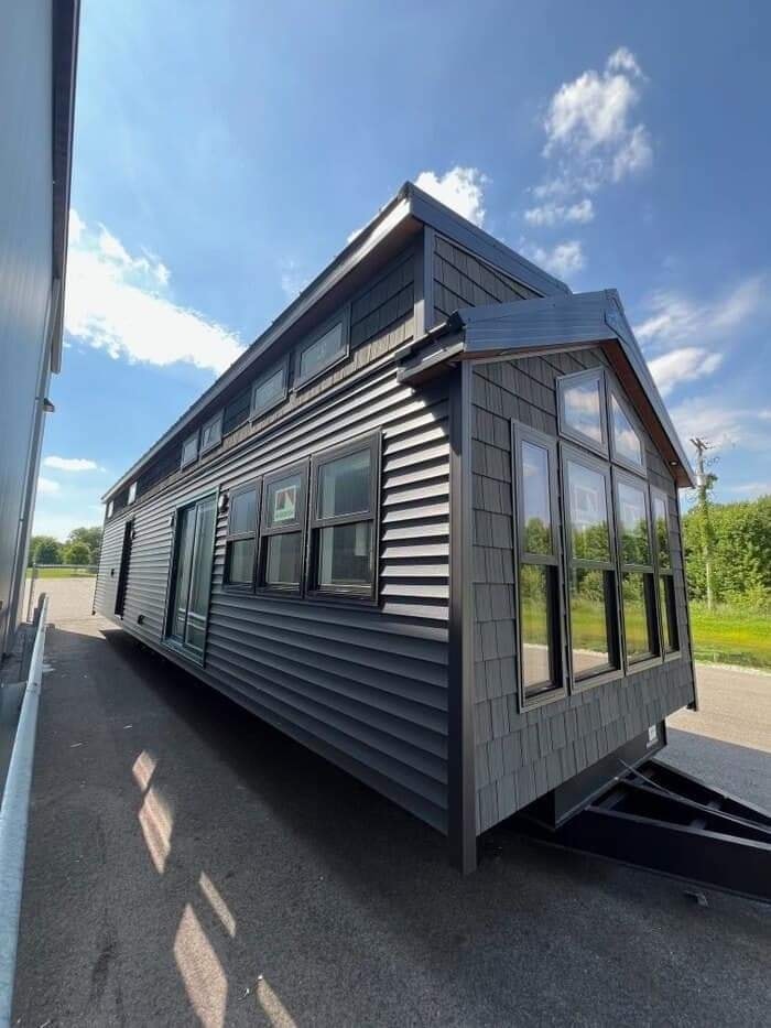 A modern tiny house on wheels with dark siding and tall windows sits on a paved area under a bright blue sky. The scene is calm and sunny.