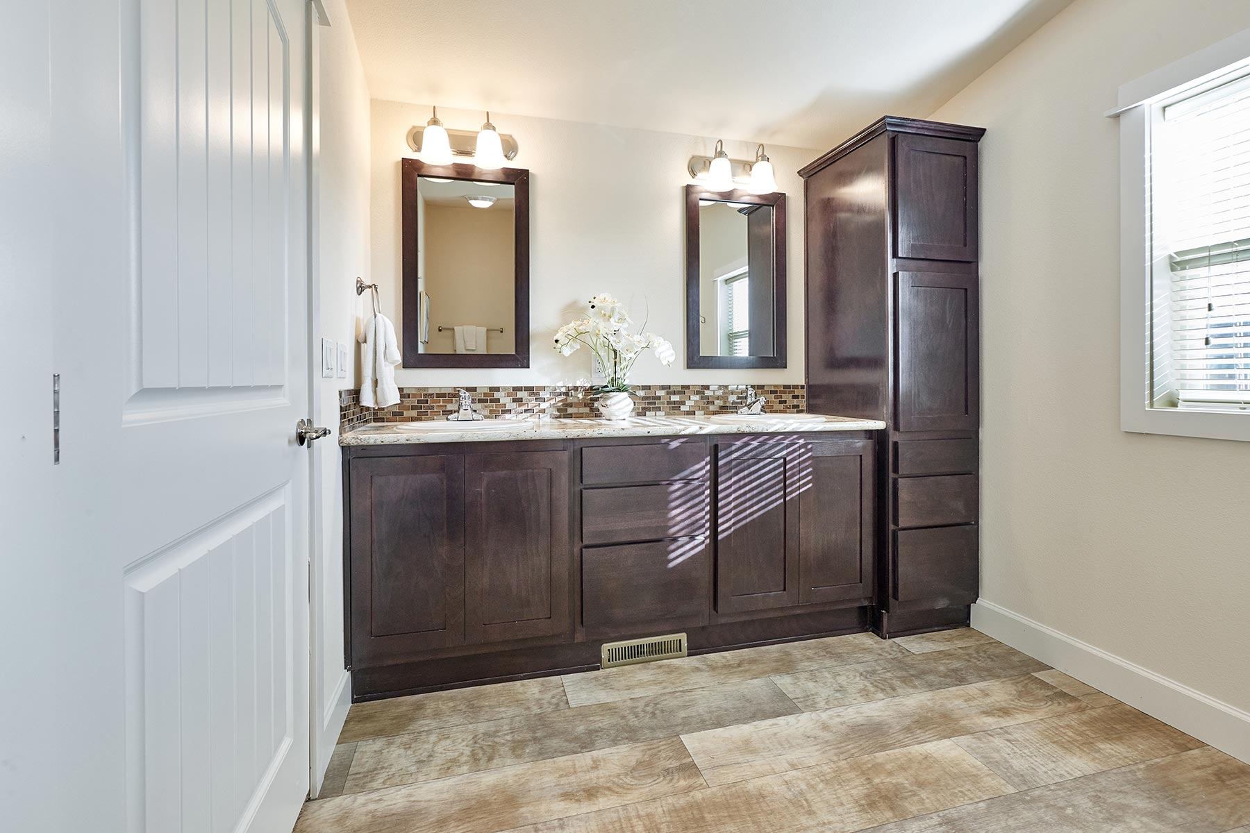 A modern bathroom with dark wood cabinets, two mirrors, and elegant light fixtures. Sunlight filters in, highlighting a vase with white flowers.