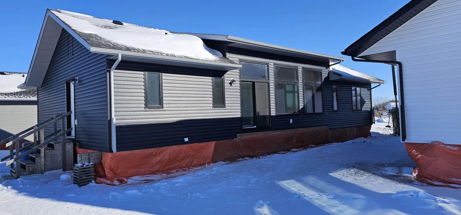 A modern gray house with snow on the roof and ground. It has large windows and a red protective covering at the base. The sky is clear and blue.