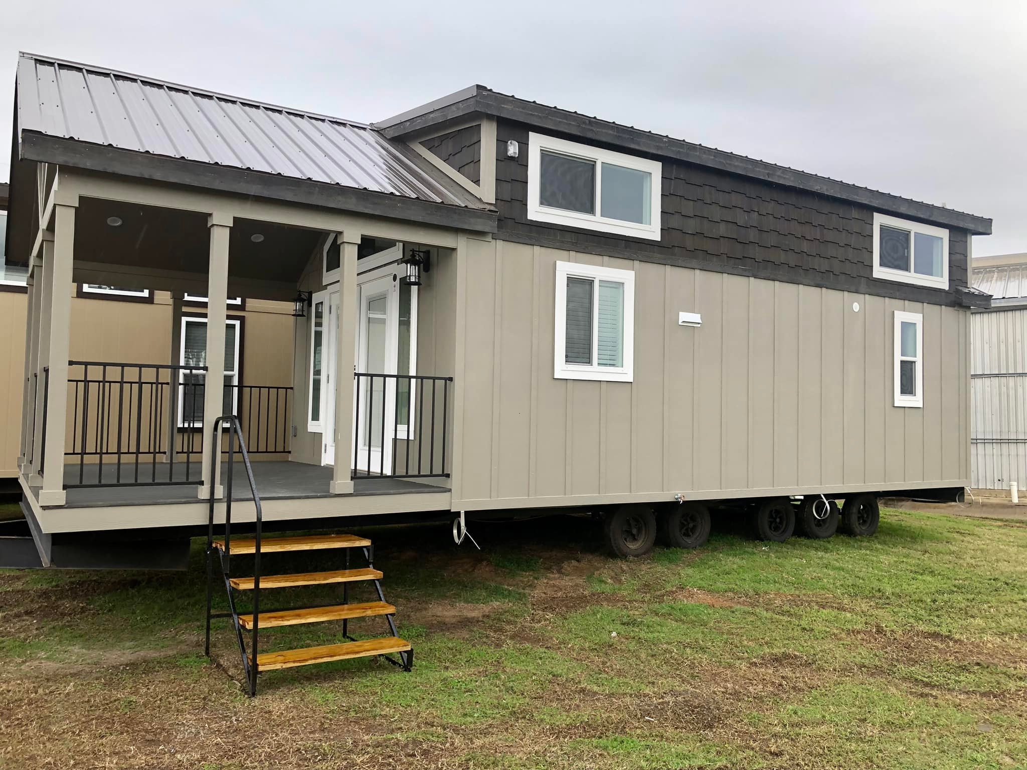 Modern tiny house on wheels with a covered porch, gray siding, and large windows. It rests on grass under an overcast sky, conveying simplicity and mobility.