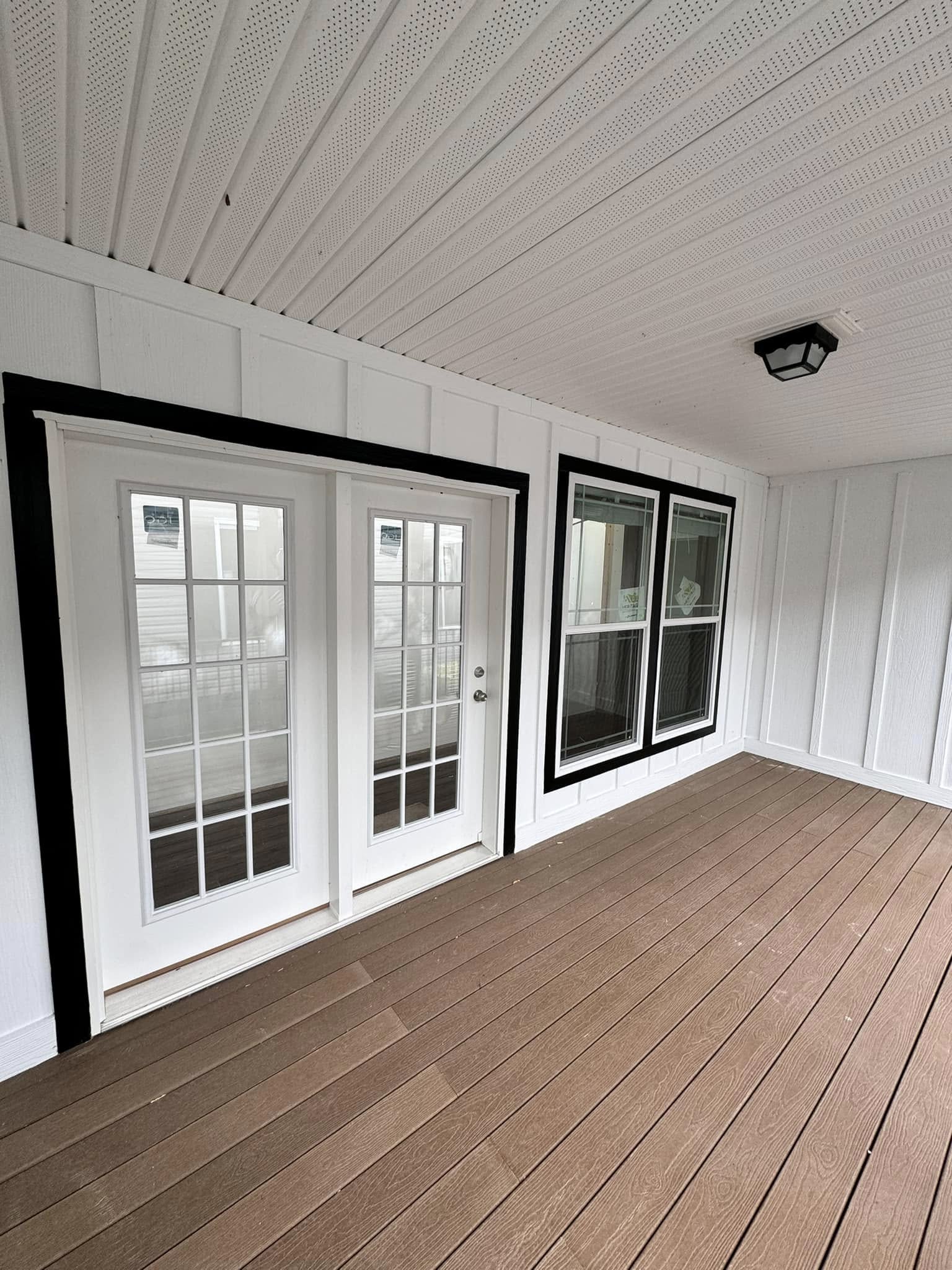 Covered porch with brown wooden flooring and white paneled walls. Features double glass doors and large windows with black frames, creating a modern, airy feel.