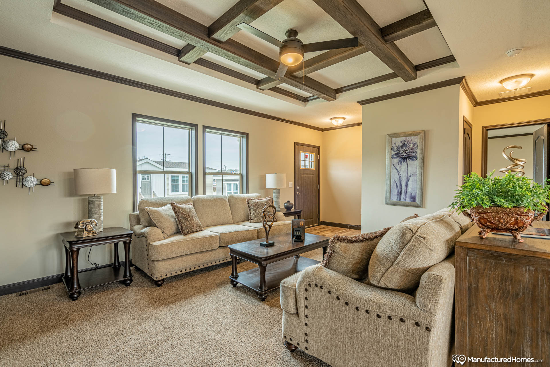 Cozy living room with beige sofas, a dark wood coffee table, and modern decor. Bright window light and a ceiling fan create a warm, inviting atmosphere.