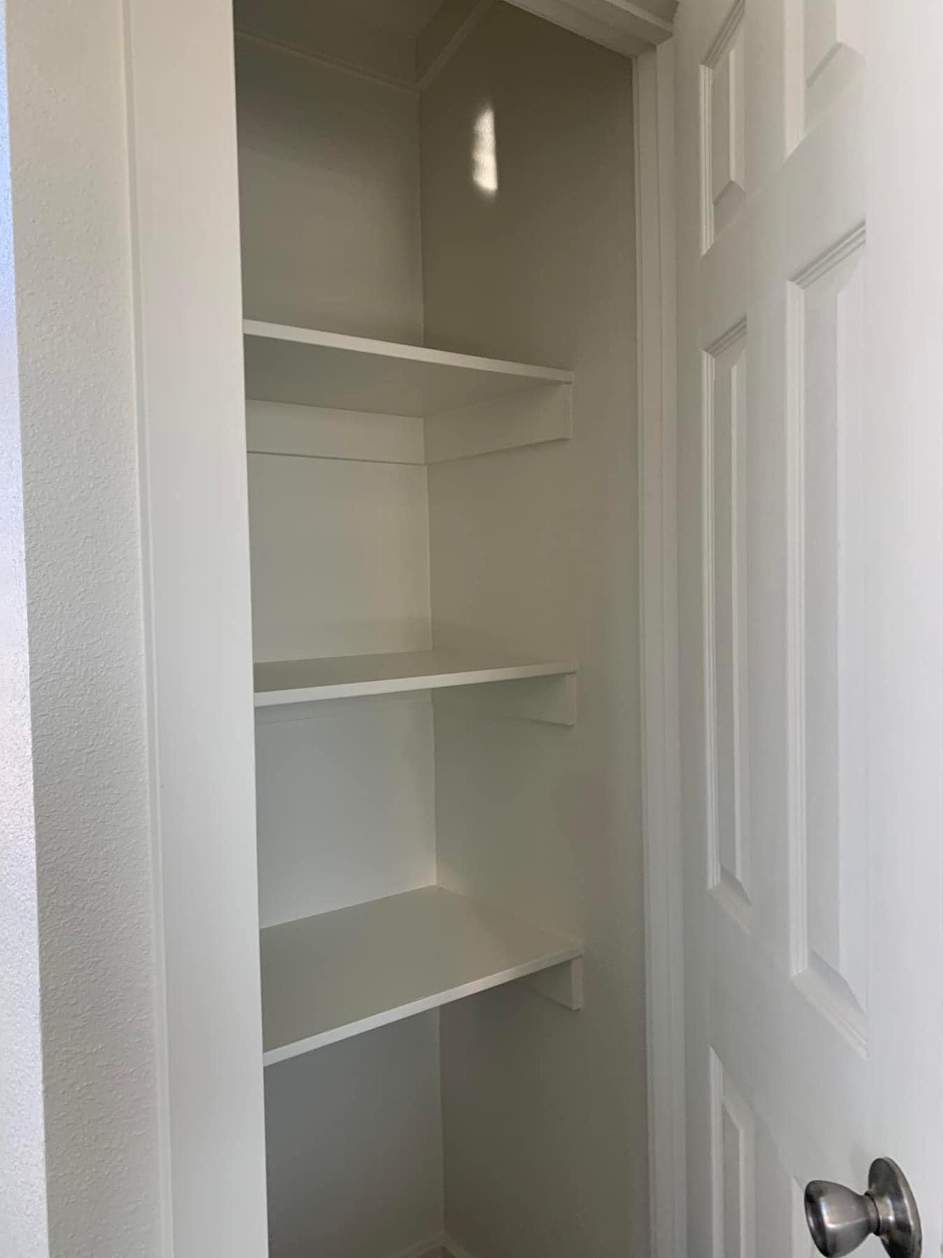 A small, empty, narrow closet with three white shelves and a white door slightly open, creating a minimal and clean tone.
