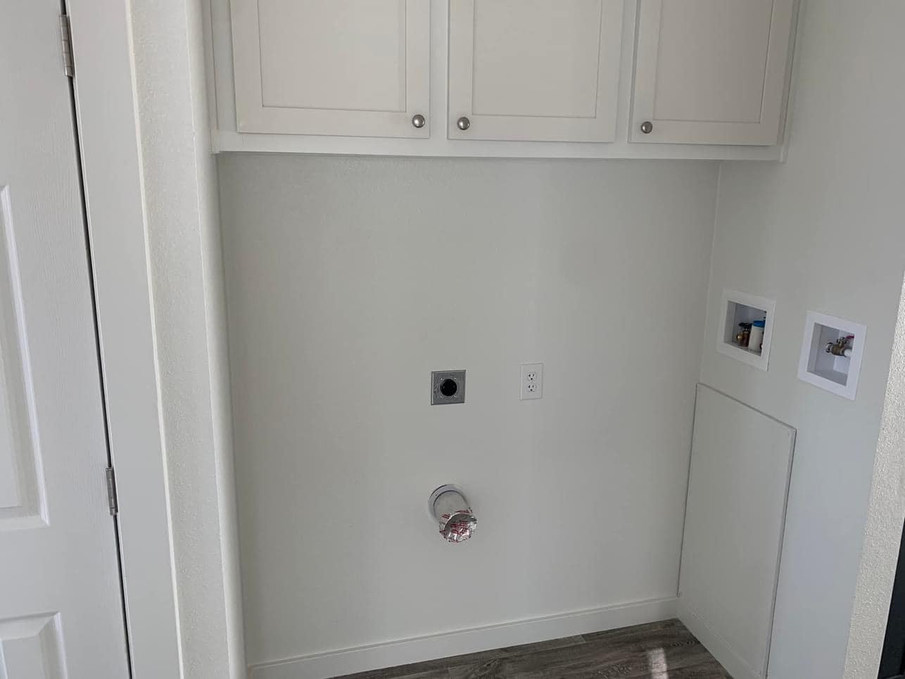 Laundry area with white cabinets, electrical outlets, and water hookups on a light-colored wall. Wooden floor, minimalistic design, calm ambiance.