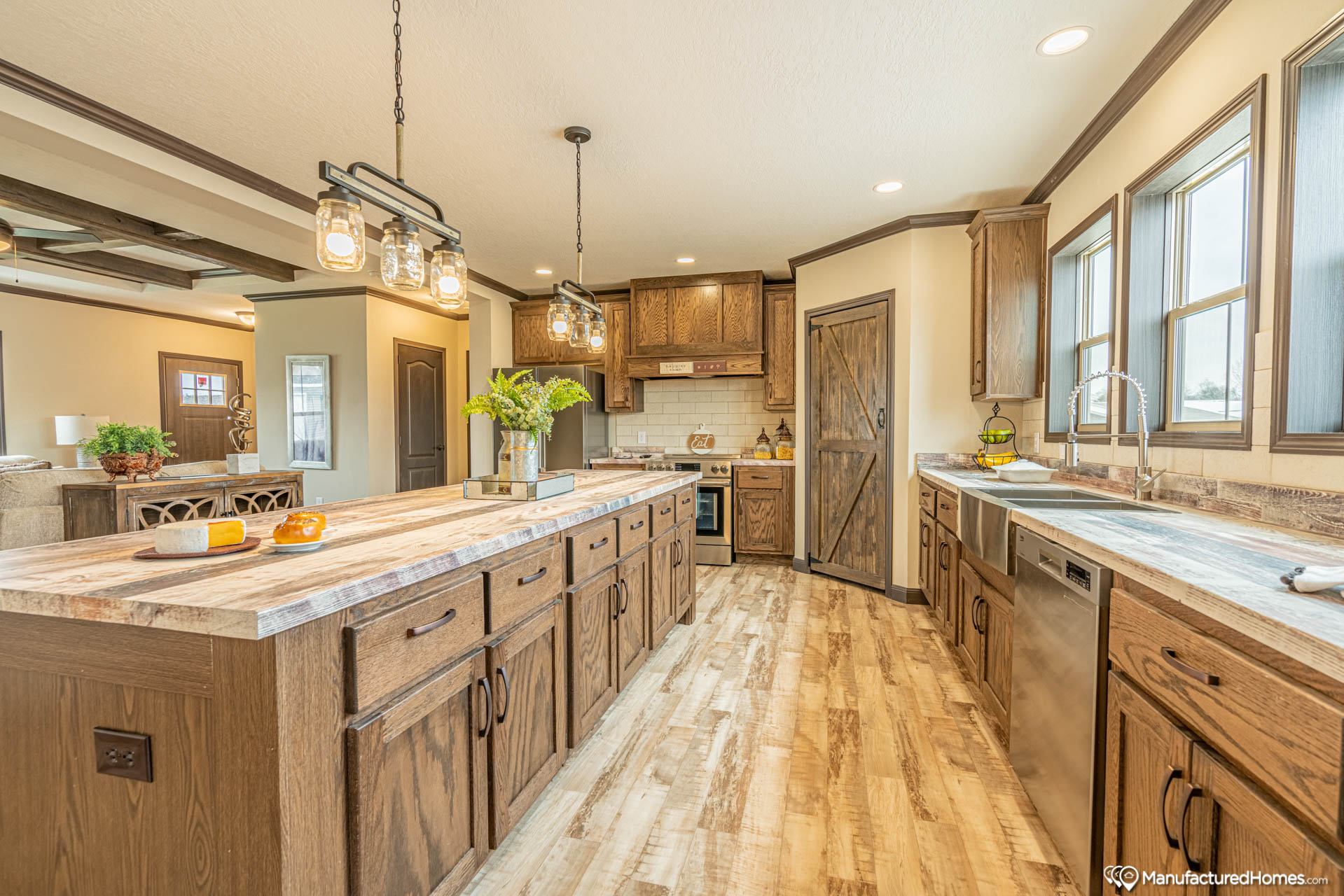 Warm, rustic kitchen with wooden cabinets and marble countertops. Pendant lights hang over the island. Large windows illuminate the space.