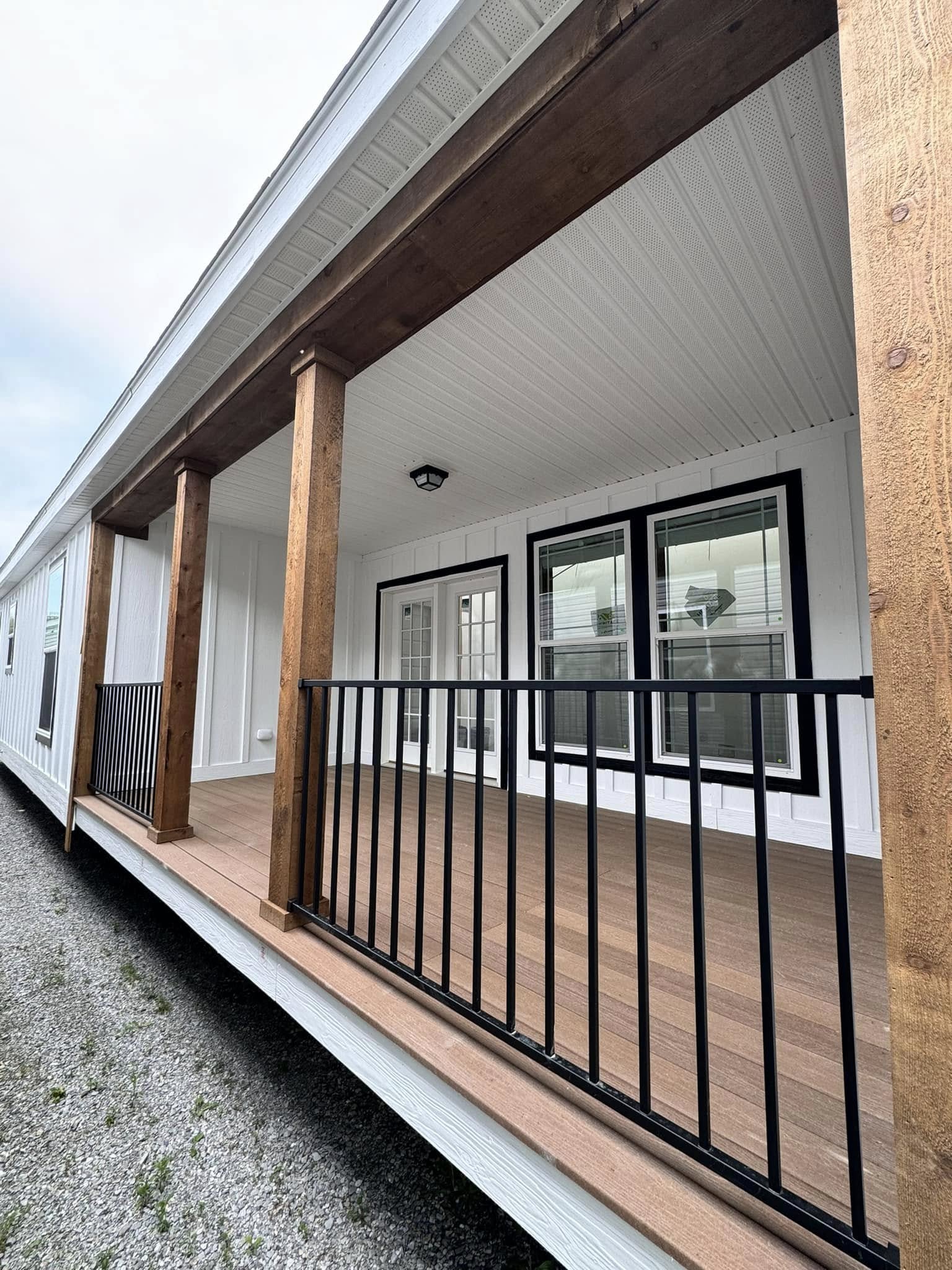 A modern porch with wood accents and black railings on a prefabricated home. It features large windows and French doors, creating a welcoming feel.