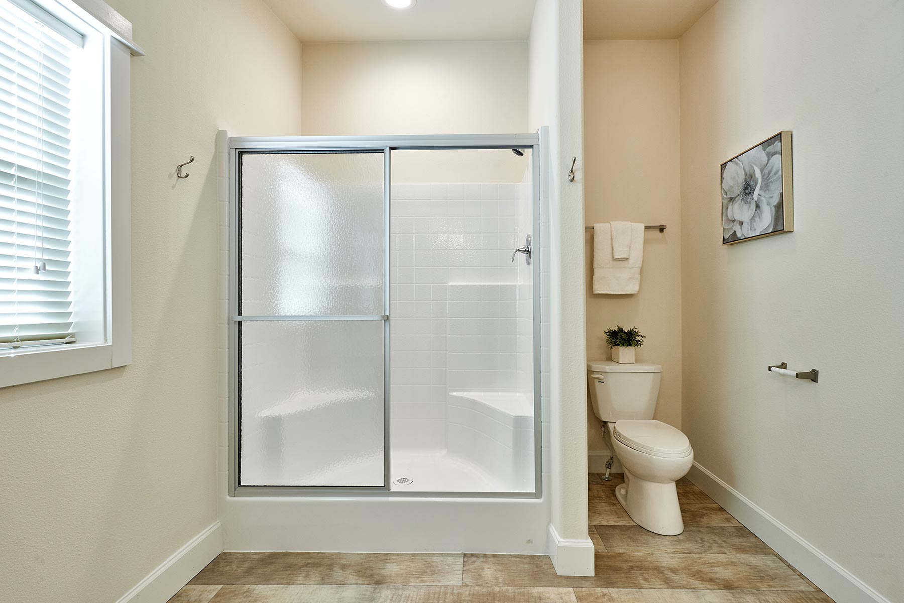 Bathroom with a frosted glass shower, white tiles, and a toilet. Light wood flooring, towel on the rack, plant on the toilet, and floral art on the wall.