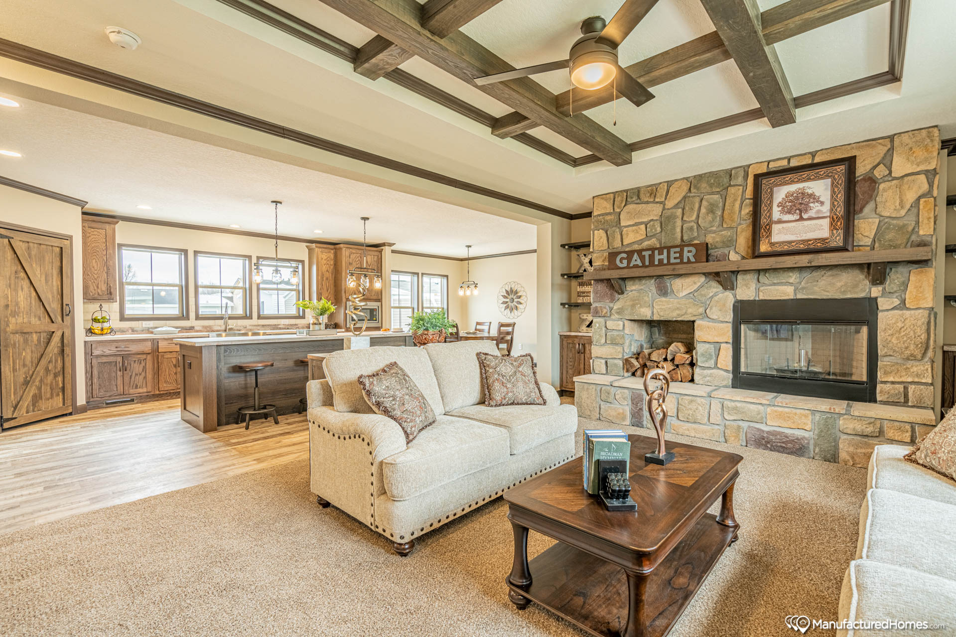 Cozy living room with beige sofas, stone fireplace, and wooden coffee table. Rustic kitchen and dining area visible, with warm lighting and wooden beams.