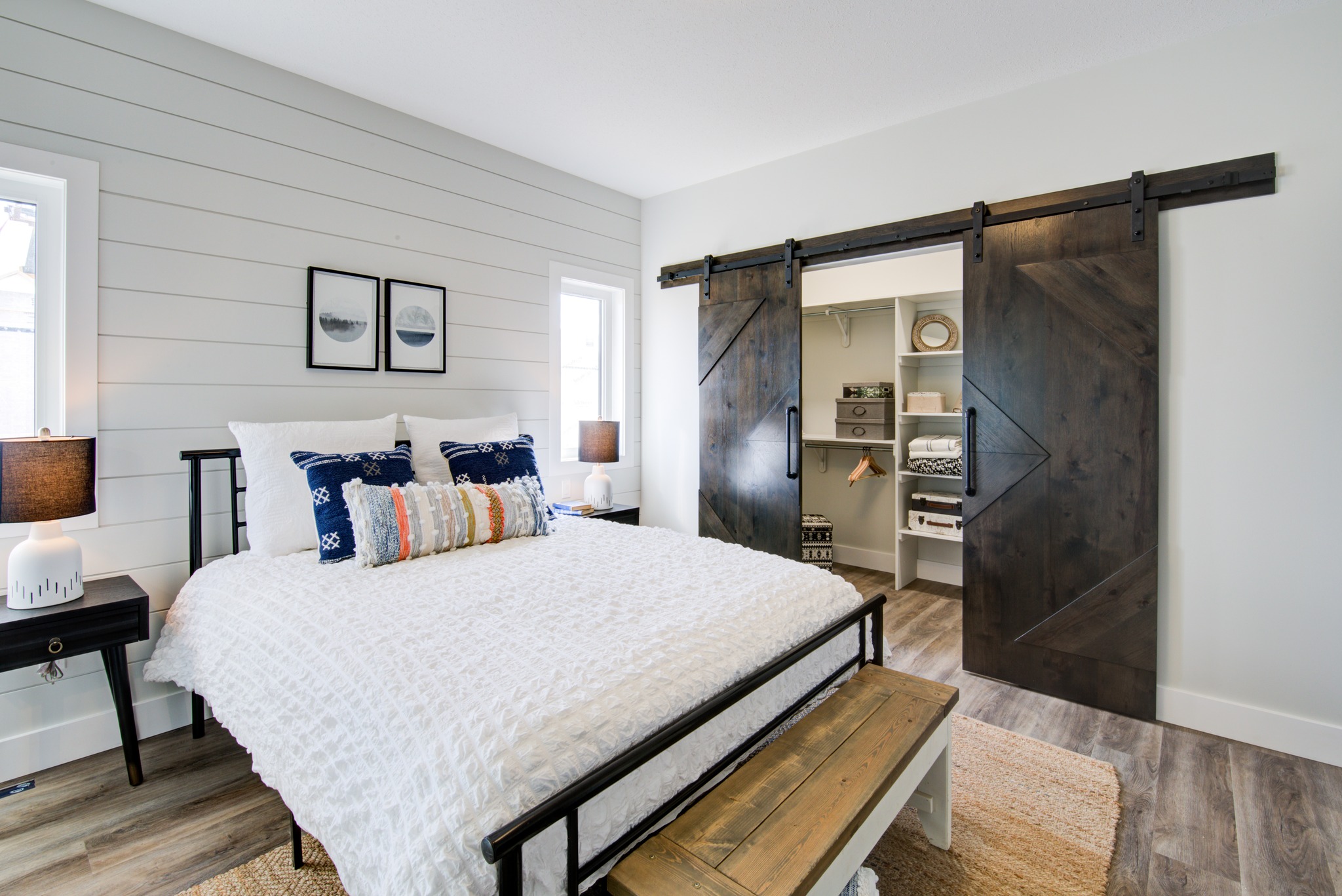 Modern bedroom with a cozy white bed, patterned pillows, and dark wood sliding barn doors revealing a neatly organized closet. Warm, inviting atmosphere.