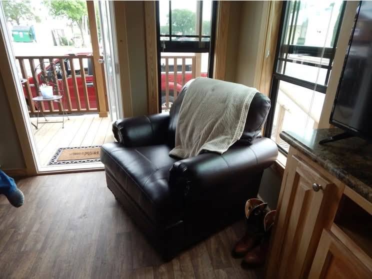 Cozy living room with a leather armchair draped with a blanket, rustic hardwood floor, cowboy boots, and an open door leading to a porch. Tranquil and inviting.