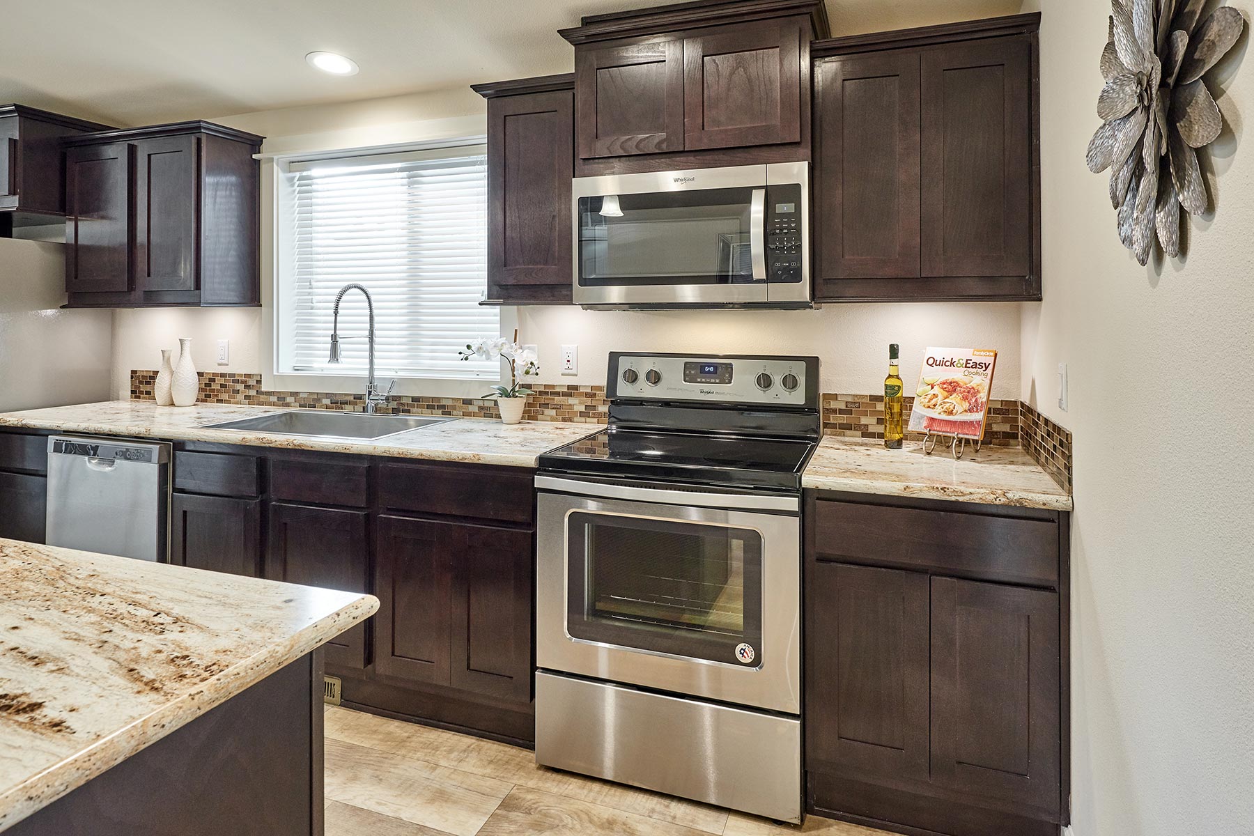 Modern kitchen with dark wooden cabinets and stainless steel appliances, including an oven and microwave. A cookbook and olive oil are on the counter.