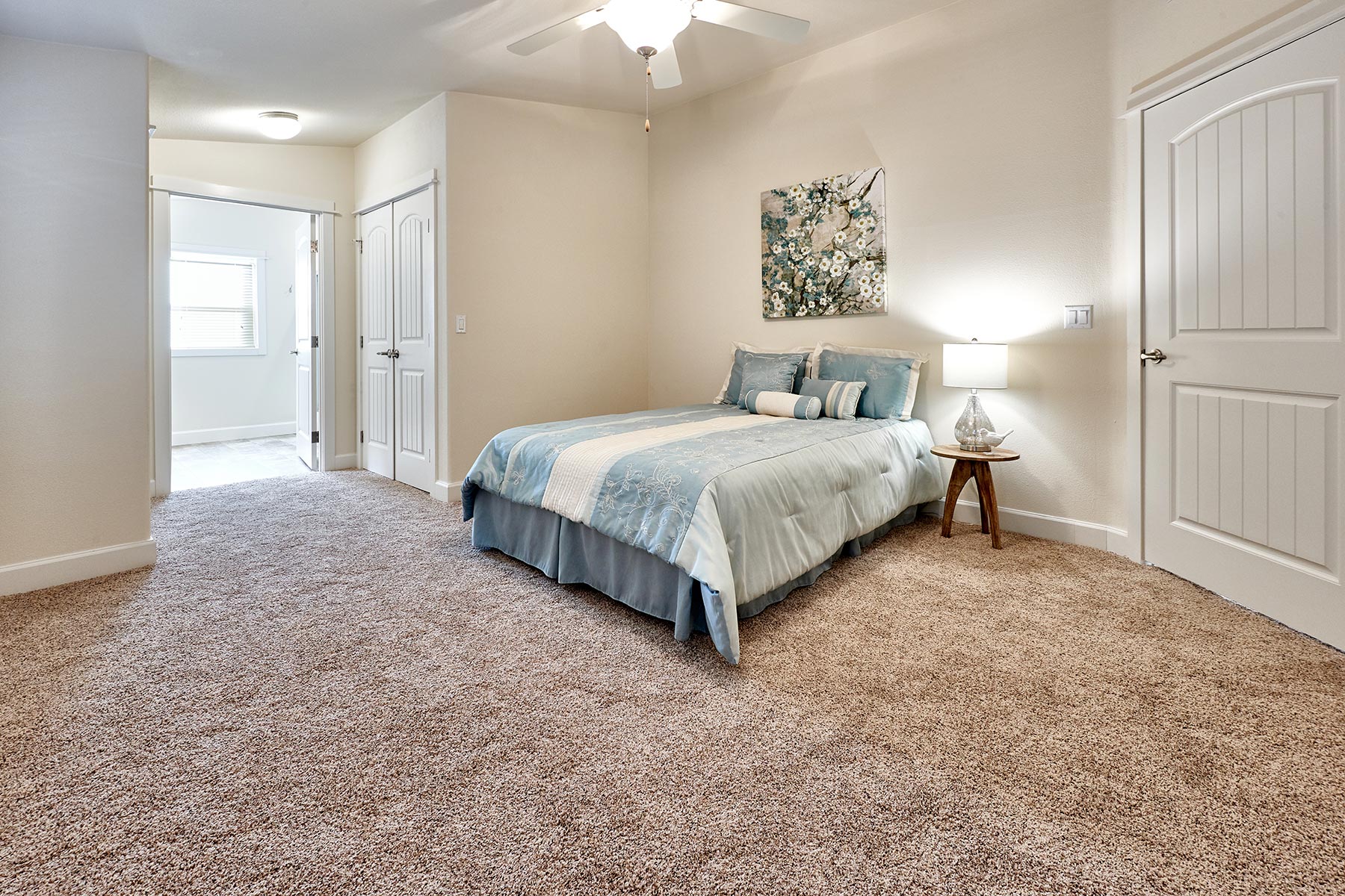A cozy bedroom with beige walls and carpet, featuring a queen-sized bed with blue and white bedding. A small wooden nightstand holds a lit lamp, under a floral artwork.