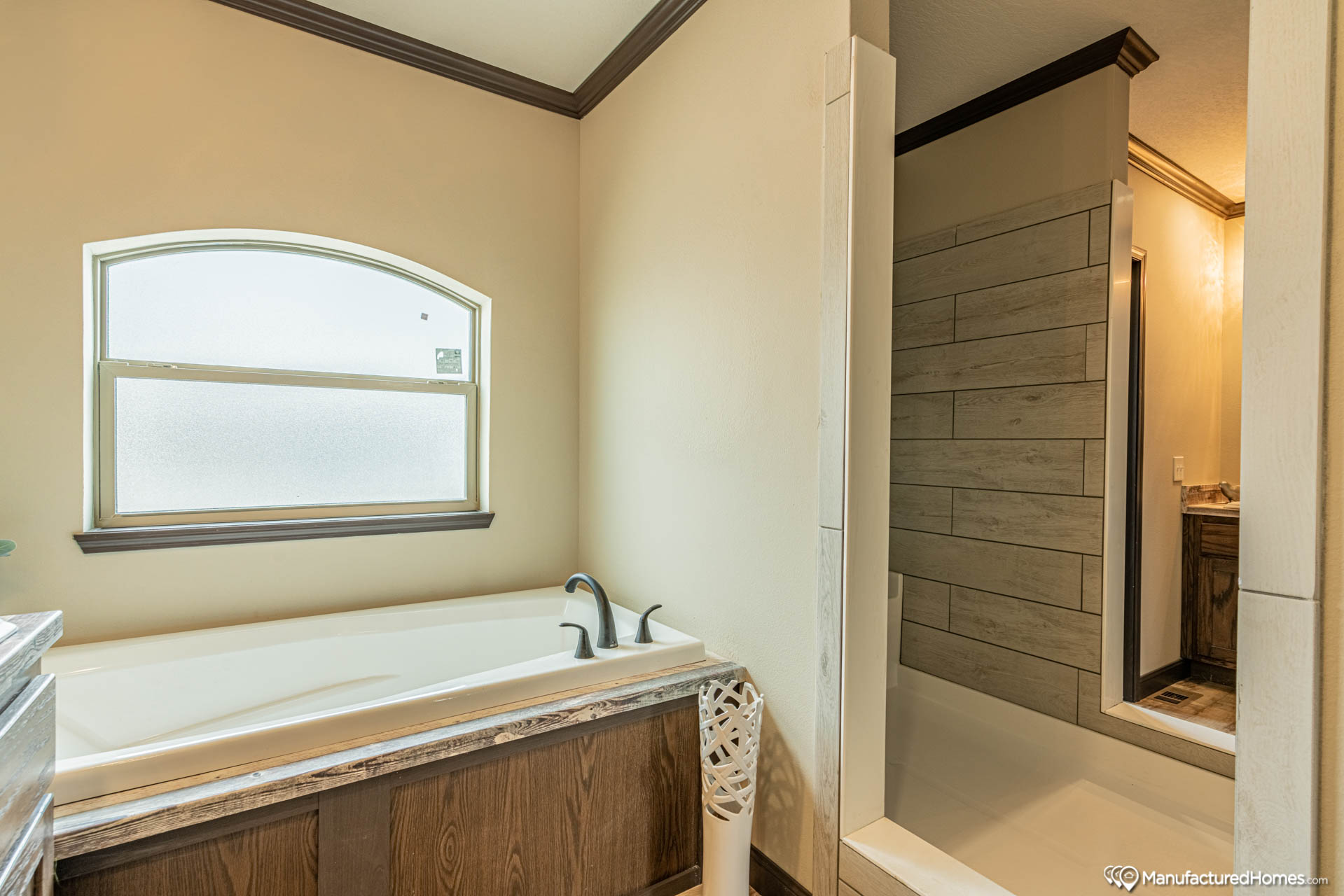 A modern bathroom with a white tub beneath a frosted arched window, wood accents, and a stone-tiled walk-in shower, creating a calm and relaxing space.